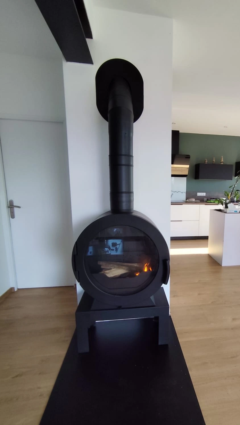 Poele a bois cylindrique noir avec vitre ronde et flambee, sur socle metallique dans un sejour ouvert sur une cuisine
