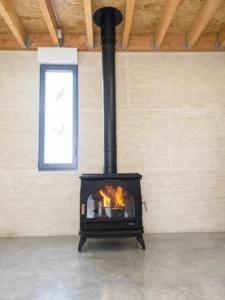 Poele a bois en fonte noir avec flambee, sur sol beton devant un mur en chanvre, sous des poutres en bois